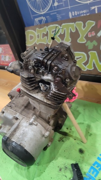 Honda ATC 70 engine sitting on a green workbench surface, 'DIRTY...' sign visible in background
