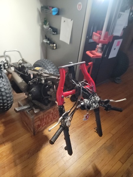 Honda ATC 70 red frame stripped bare in a living room, engine sitting off to the side on a wooden crate