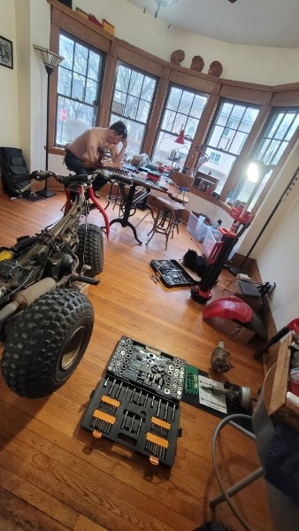 Shirtless person working on a Honda ATC in a living room with bay windows, tools and socket sets spread across the hardwood floor