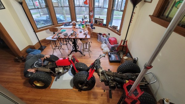 Overhead view of a living room converted into a workshop with two Honda ATCs, tools, and socket sets spread across hardwood floors beneath bay windows
