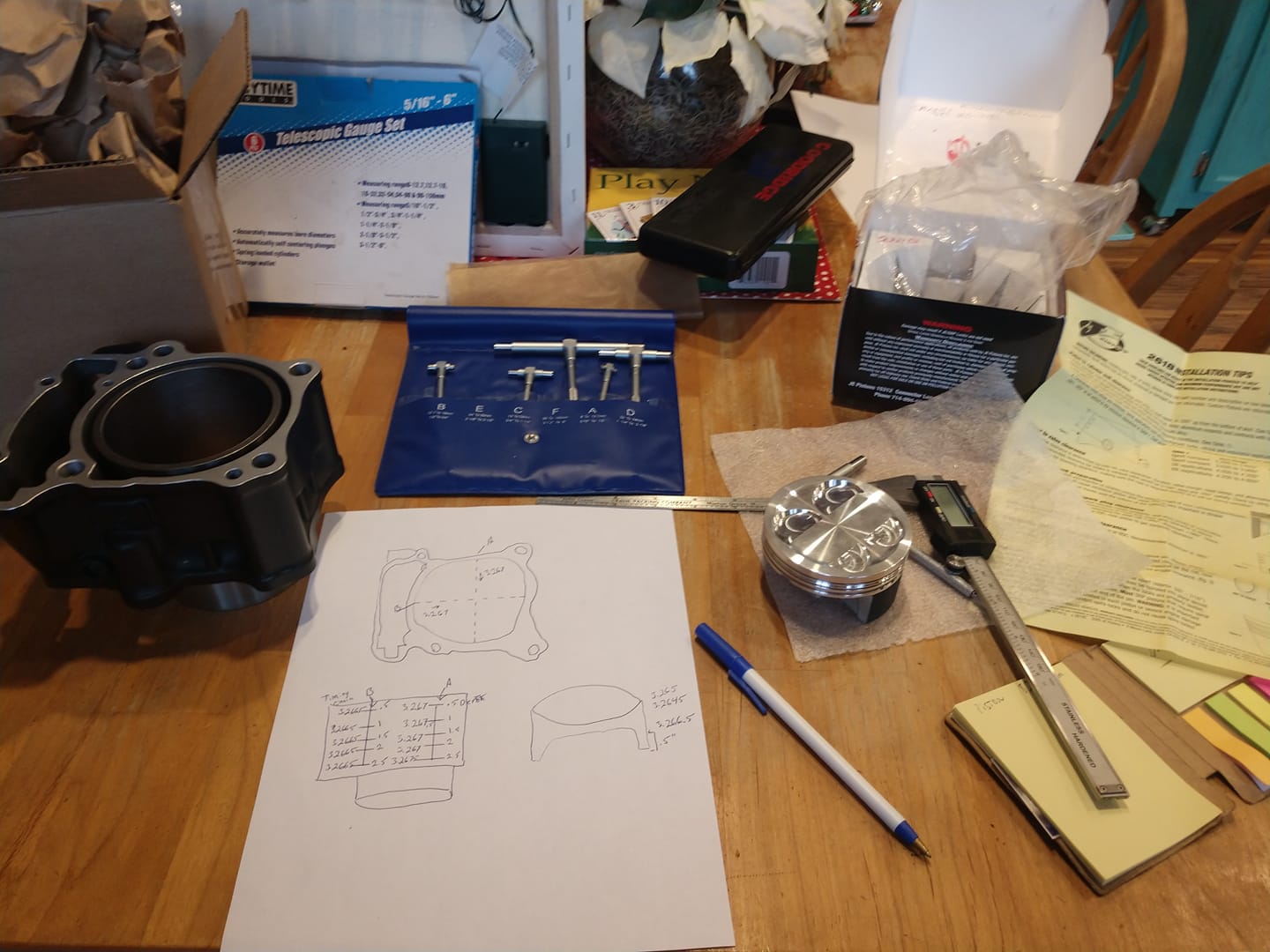CBR300R engine parts on the kitchen table — cylinder jug, piston, telescopic gauge set, digital calipers, spec sheets, and handwritten notes