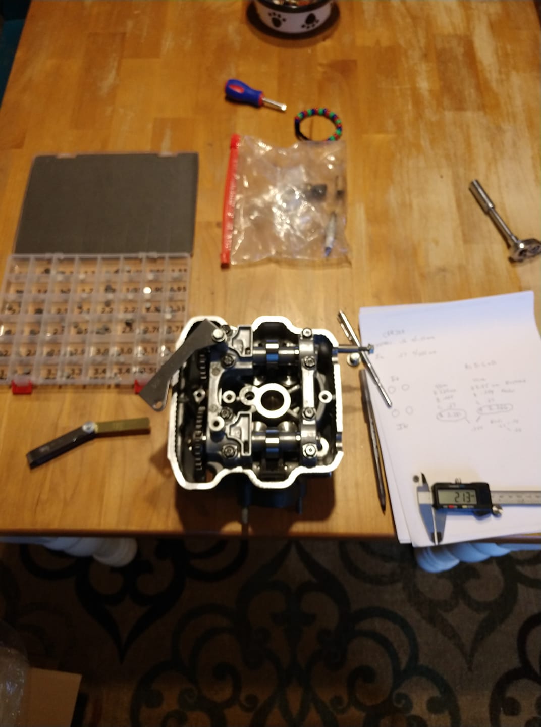 CBR300R cylinder head with cams installed on the kitchen table — shim tray, telescopic gauges, calipers, and handwritten valve clearance calculations on paper