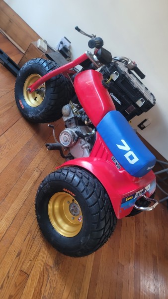 Completed Honda ATC 70 restoration — red and blue bodywork, gold wheels, ProTaper bars, ATC 70 decals, sitting on hardwood floor
