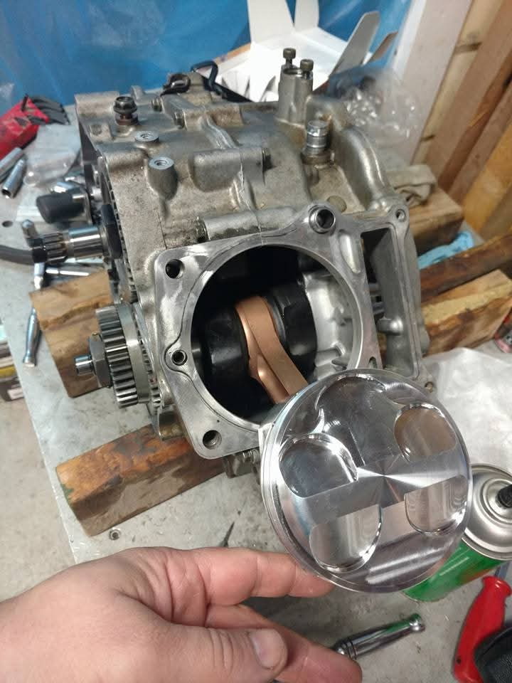 Steve holding a polished JE big-bore piston next to the open Raptor 700 cylinder, showing the oversized piston dome relative to the bore