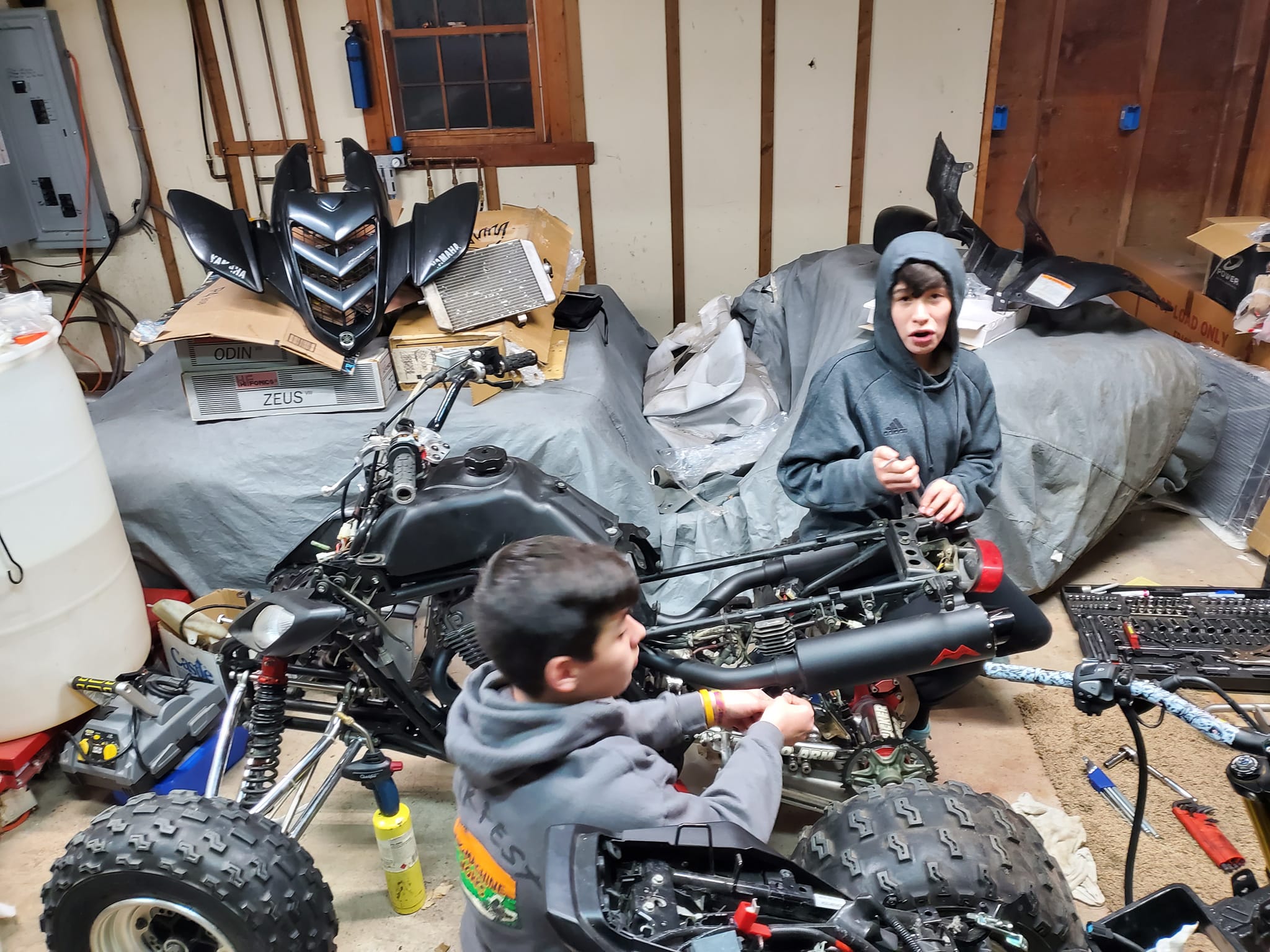 Two kids working on a Yamaha Raptor 700 in Steve's garage — one using a wrench on the engine area, the other holding parts, bodywork removed